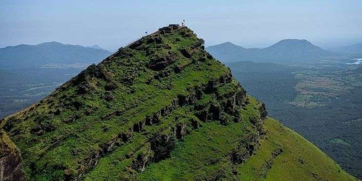 A man stands on a mountain peak overlooking a lush green grassy area.