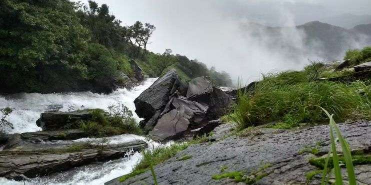 A scenic waterfall in the mountains surrounded by rocks and grass, part of a challenging trek to Ballalarayana Durga Fort.