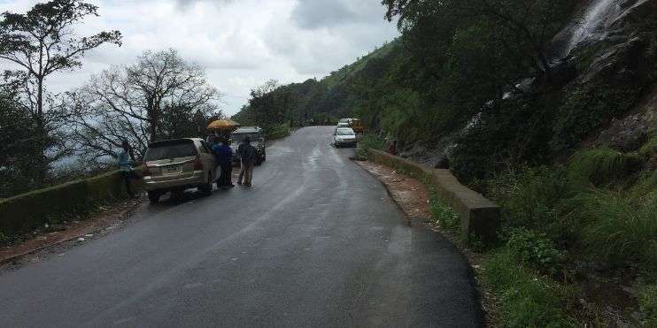 A scenic road in Charmadi Ghat with a car and two people walking, surrounded by lush valleys, waterfalls, and forests.