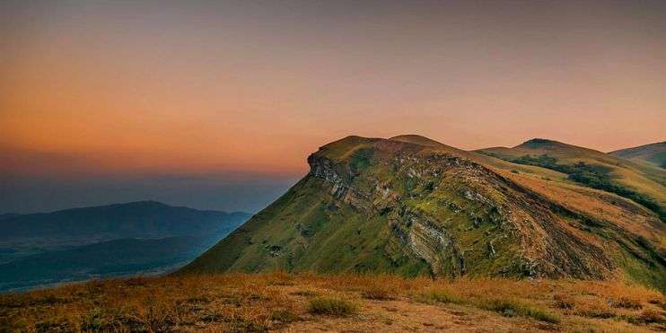 Sun setting over lush green mountain range with grass and hills in Kemmangundi, known for gardens, waterfalls, and trekking trails.