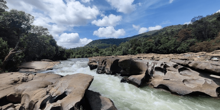 A serene river flows through rocky terrain, surrounded by trees, highlighting the sacred beauty of Amba Teertha in Kalasa.