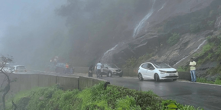 A car travels along a scenic road with a stunning waterfall cascading in the background at Charmadi Ghat near Kalasa.