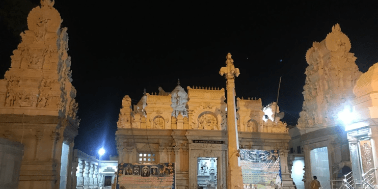 A large white Shri Veerabhadra Swamy Temple illuminated at night, surrounded by numerous statues, creating a serene atmosphere.