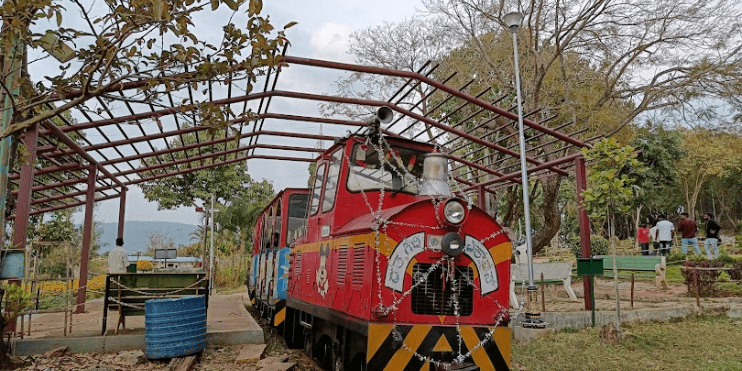 A red toy train is parked in Mahatma Gandhi Park, inviting visitors to enjoy a scenic ride through its beautiful landscape.