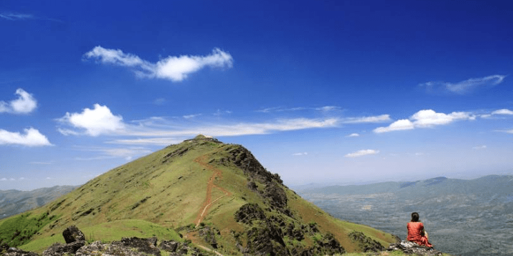 A man sits atop Mullayanagiri Peak, enjoying a panoramic view under a clear blue sky, perfect for adventure enthusiasts.