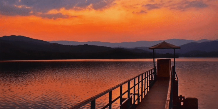 A serene sunset over Ayyanakere Lake, with a pier in the foreground and majestic mountains silhouetted in the background.