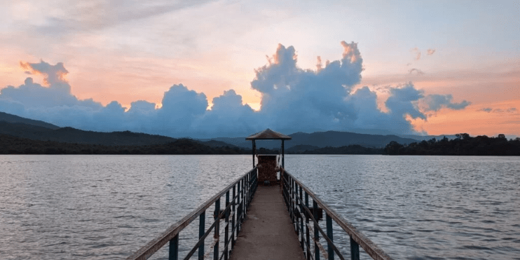 A serene pier at sunset, with vibrant clouds reflecting on the tranquil waters of Ayyanakere Lake, surrounded by lush greenery.