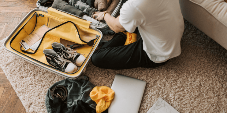 A man sits on the floor with a suitcase and laptop, preparing for a trip with essential packing items nearby.