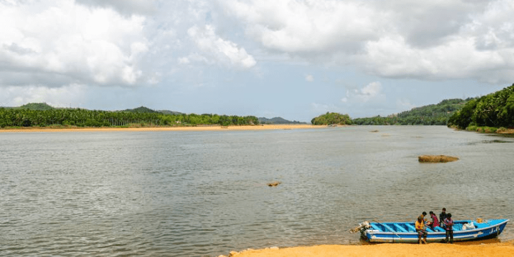 A boat carrying people glides along the serene Netravathi River, surrounded by lush forests and rolling hills.