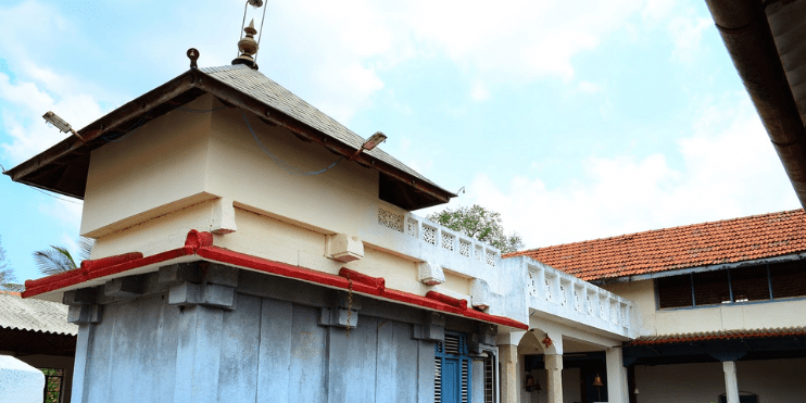 A small temple with a red roof and white tower, surrounded by lush greenery and hills in Chikmagalur's serene landscape.