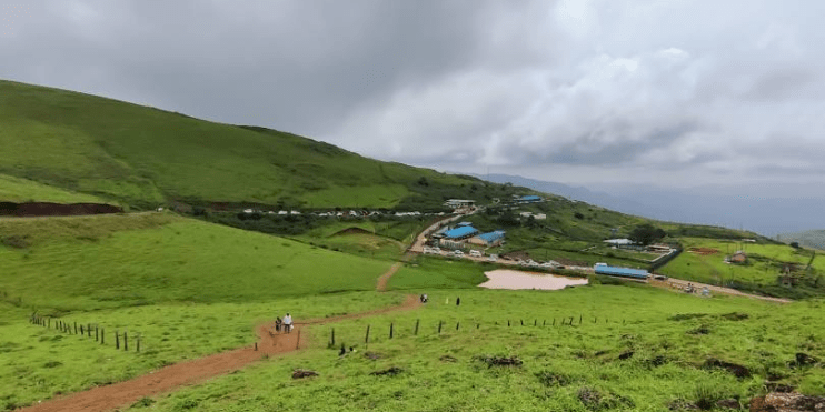 A picturesque green valley featuring a winding road and charming houses, set against the backdrop of the Baba Budangiri Hills. 