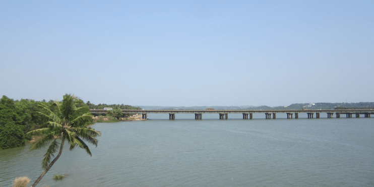 A serene bridge spans the Netravathi River, surrounded by lush greenery and smooth boulders, inviting tranquility and relaxation.