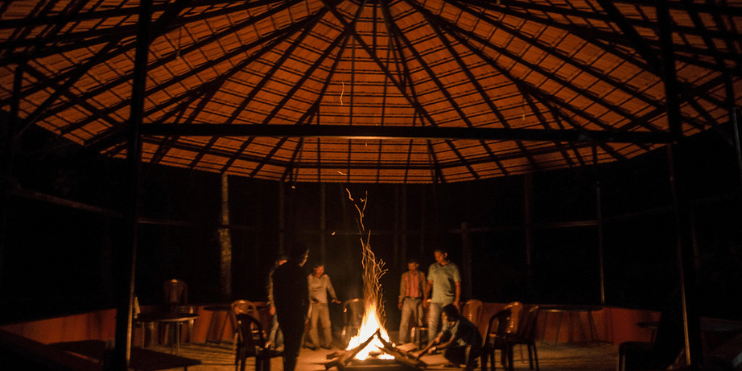 A group of people gathered around a bonfire in a cozy hut, sharing stories under a starry sky.