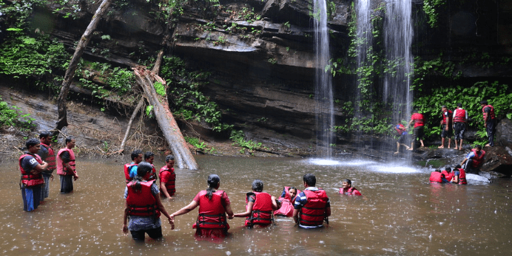 A group of individuals in life jackets stands by a river, enjoying the scenic view of a nearby waterfall.