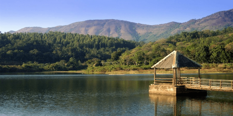 A wooden pier extends over Hirekolale Lake, framed by lush greenery and majestic mountains in a tranquil setting.