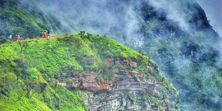 People stroll on a grassy mountain slope, with the historic Ballalarayana Durga Fort rising majestically behind them.