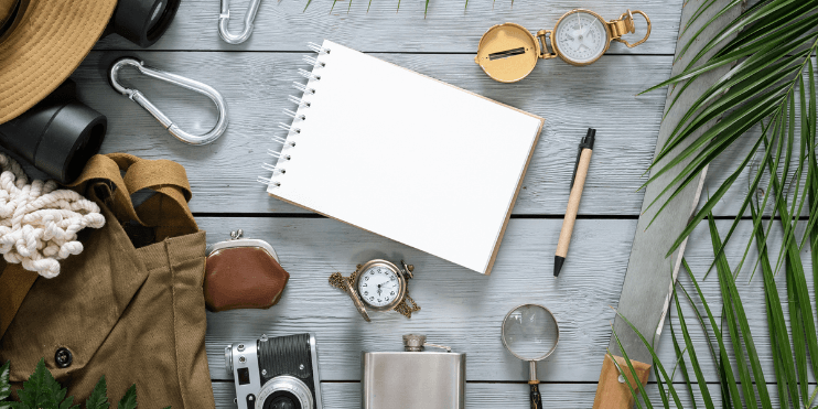 A wooden table displaying a notebook, pen, hat, camera, and various travel essentials for an organized trip.