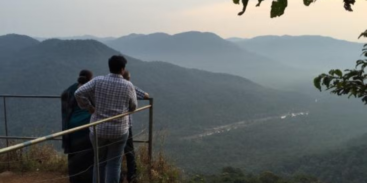 Two people stand on a railing, gazing at the mountains from Jenukal Gudda, enjoying the scenic view.