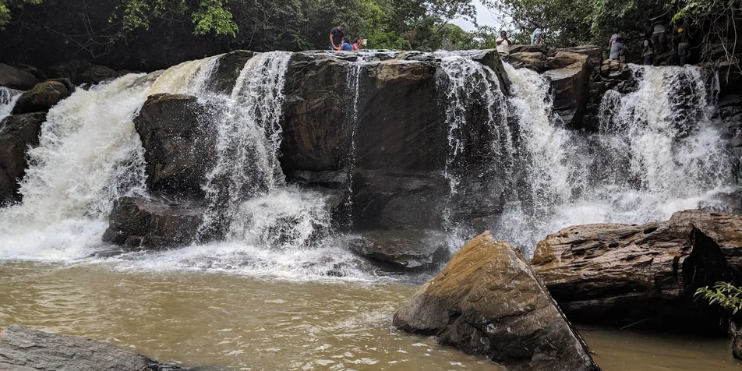A serene waterfall, Mookanamane Falls, surrounded by rocks and lush trees, perfect for a peaceful picnic in nature.